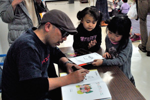 絵本にサインをするサトシンさんと見つめる子どもたちの画像